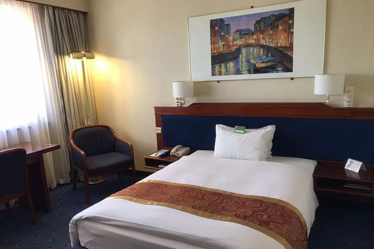 A hotel room with a large bed, a desk, a chair, a nightstand, and a painting on the wall.