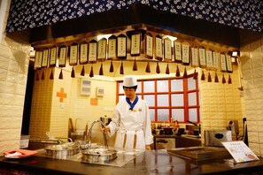 A chef in a white uniform with a blue apron standing at a stainless steel counter with various cooking utensils and dishes.
