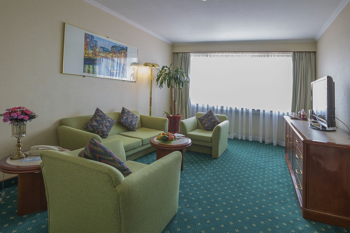 A hotel room with a green carpet, two green armchairs, a sofa, a small round table, a potted plant, a television, and a painting on the wall.