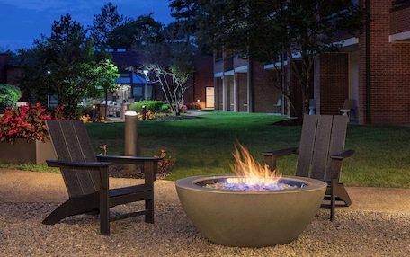 A fire pit with two Adirondack chairs in a landscaped outdoor area with a building in the background.