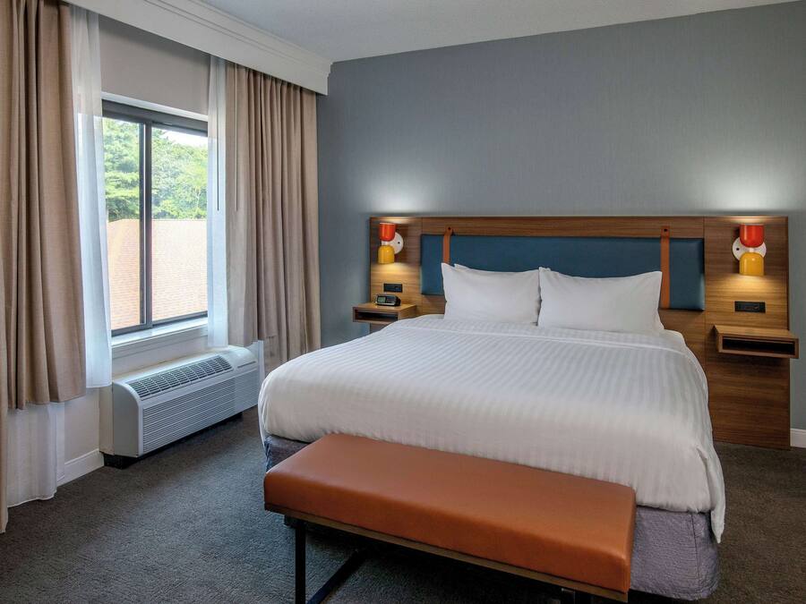 Habitación de hotel con una cama grande, un banco, una ventana con cortinas y lámparas de noche.