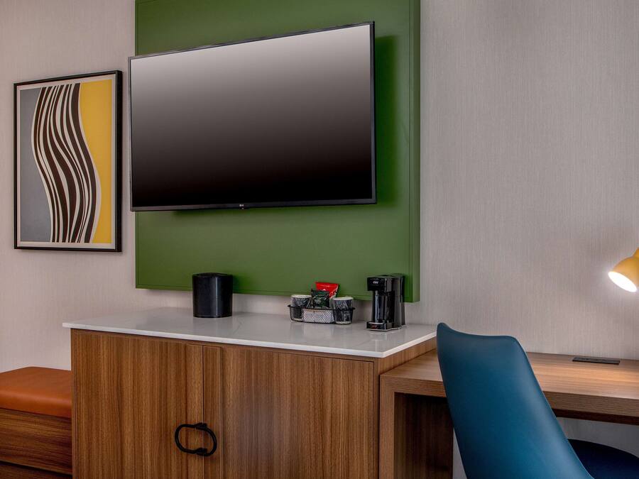 Quarto moderno com TV de tela plana presa a uma parede verde, escrivaninha de madeira com cadeira azul, cafeteira e abajur.