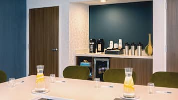 Ein Konferenzraum mit einem langen Tisch, zwei Wassergläsern, einer Kaffeestation und einem Kühlschrank.