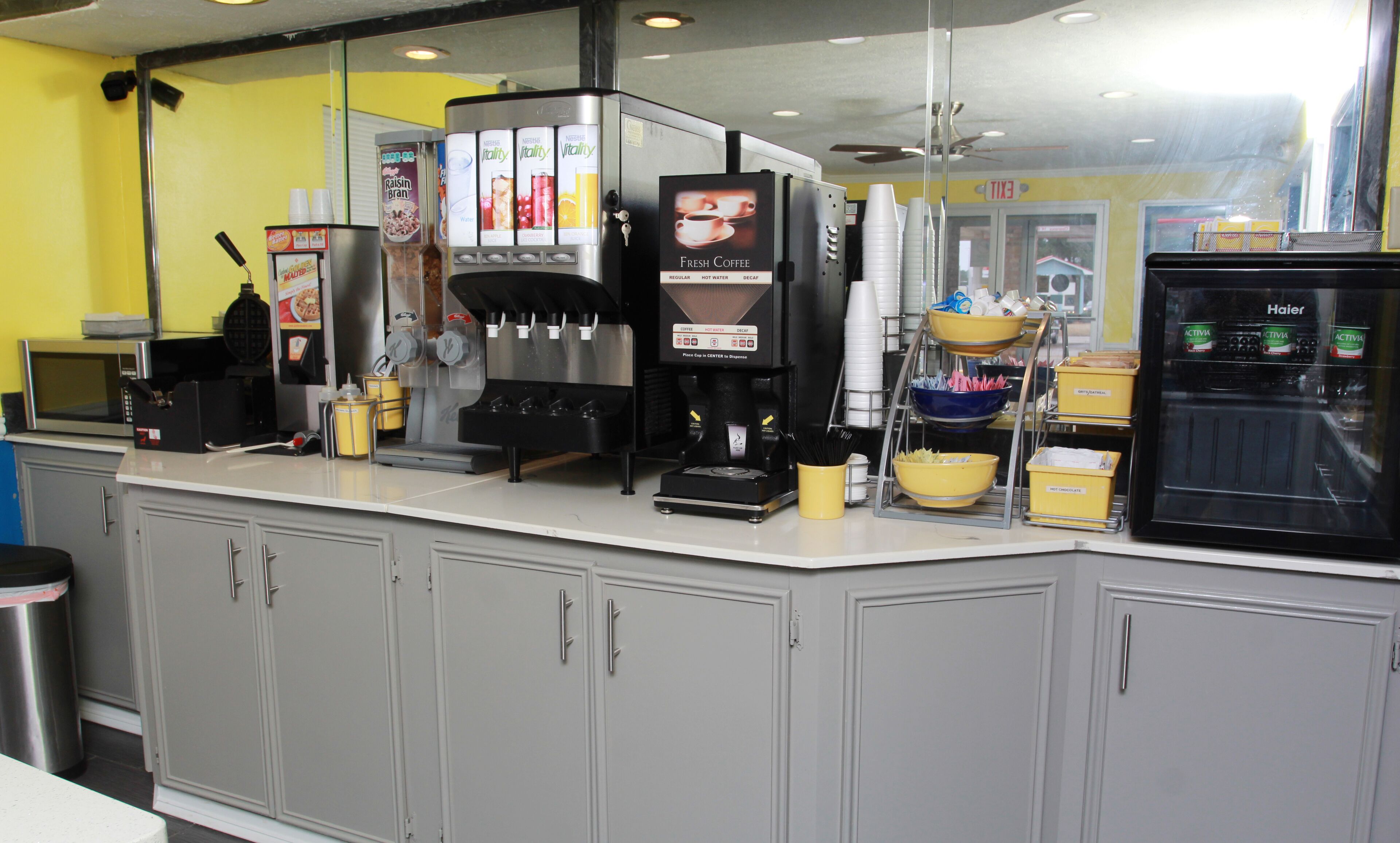 Mostrador de cafetería con máquina de café, dispensadores de condimentos y refrigerador.