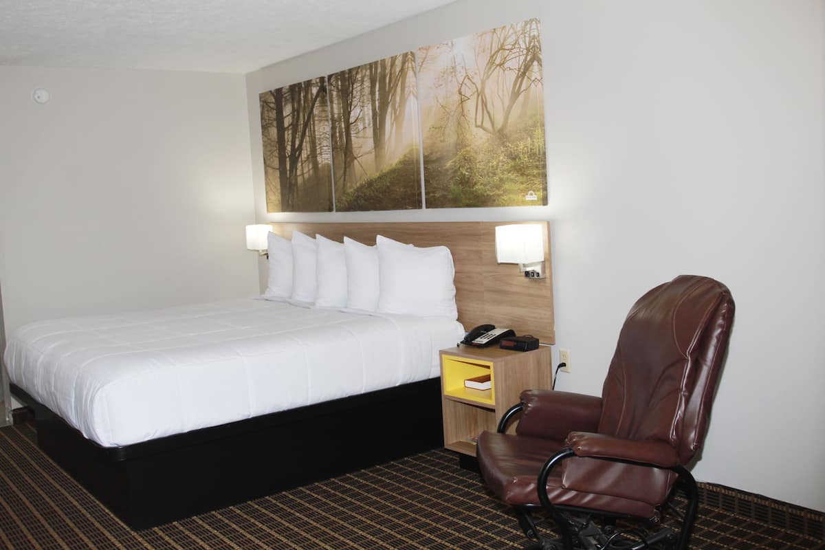 Quarto de hotel com uma cama grande, cabeceira de madeira, mesa de cabeceira com abajur, telefone e uma poltrona de couro marrom.