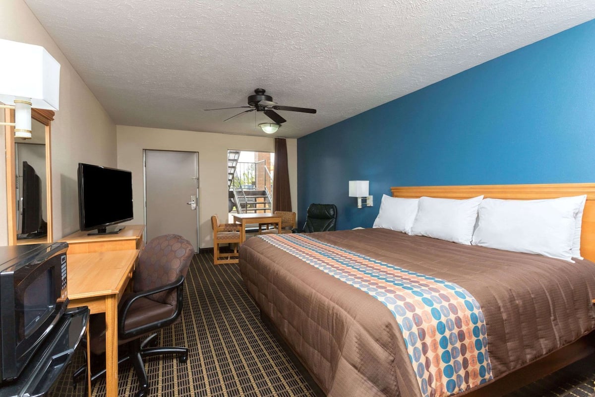 Quarto de hotel com cama, escrivaninha com televisão, uma cadeira, ventilador de teto e janela com vista.