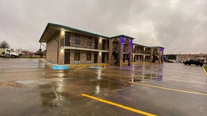 A multi-story hotel with a covered parking area and a wet ground, likely after rain. - Days Inn by Wyndham Forest (Forest)