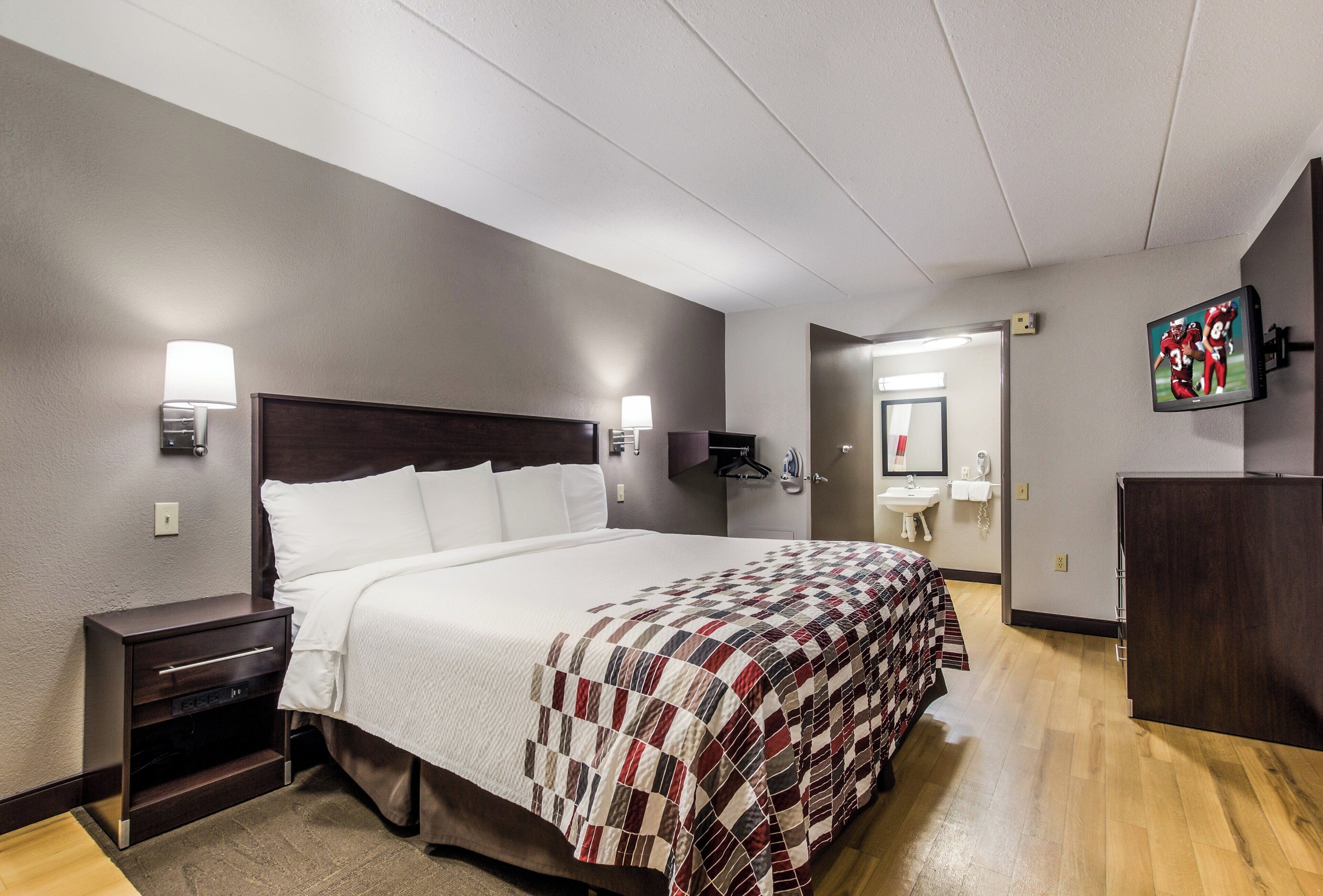 A hotel room with a bed, bedside tables, a wall-mounted TV, and a bathroom visible through an open door.