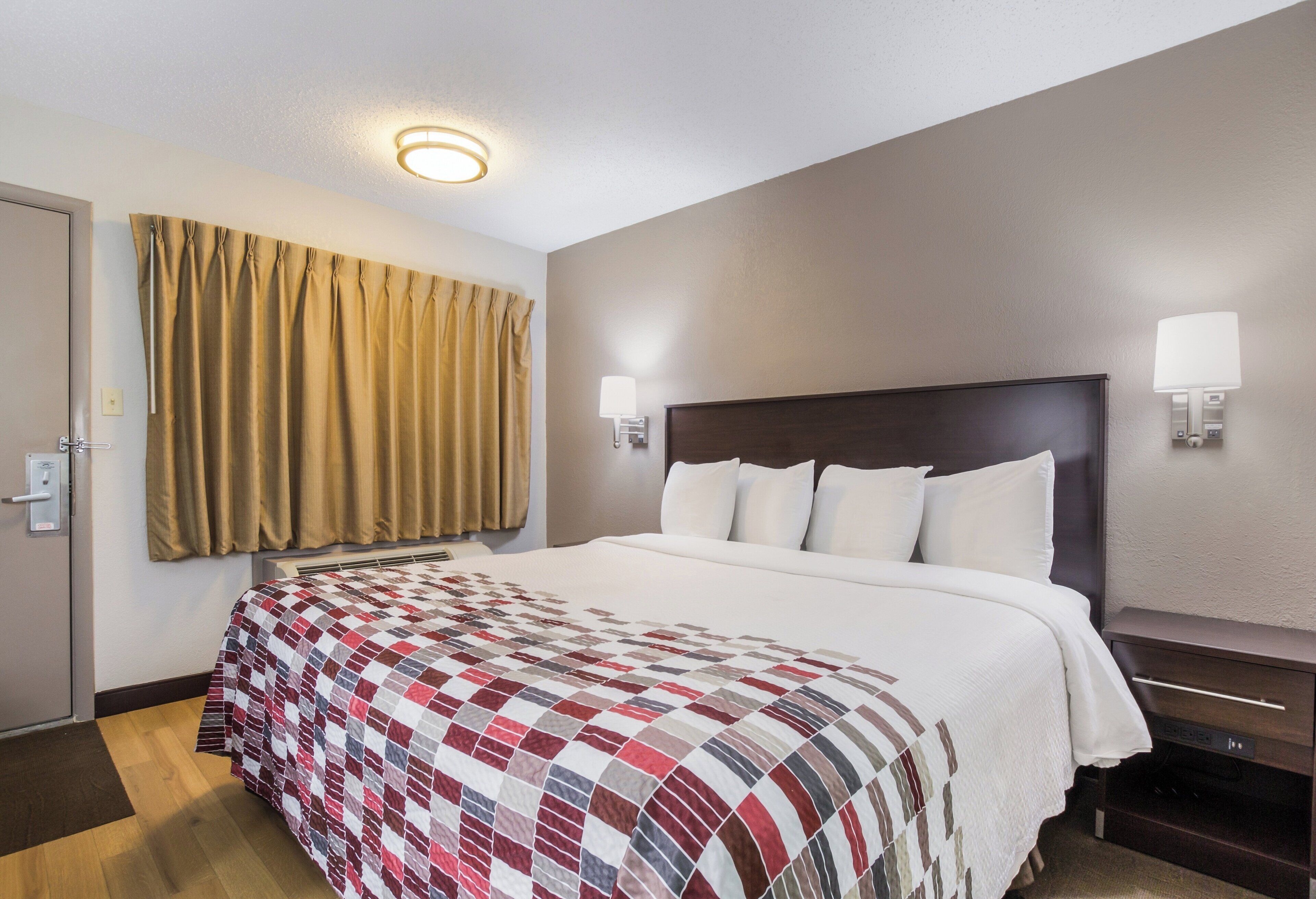 Una habitación de hotel con una cama grande, un cabecero, dos lámparas de noche, un ventanal con cortinas y una puerta con cerradura.