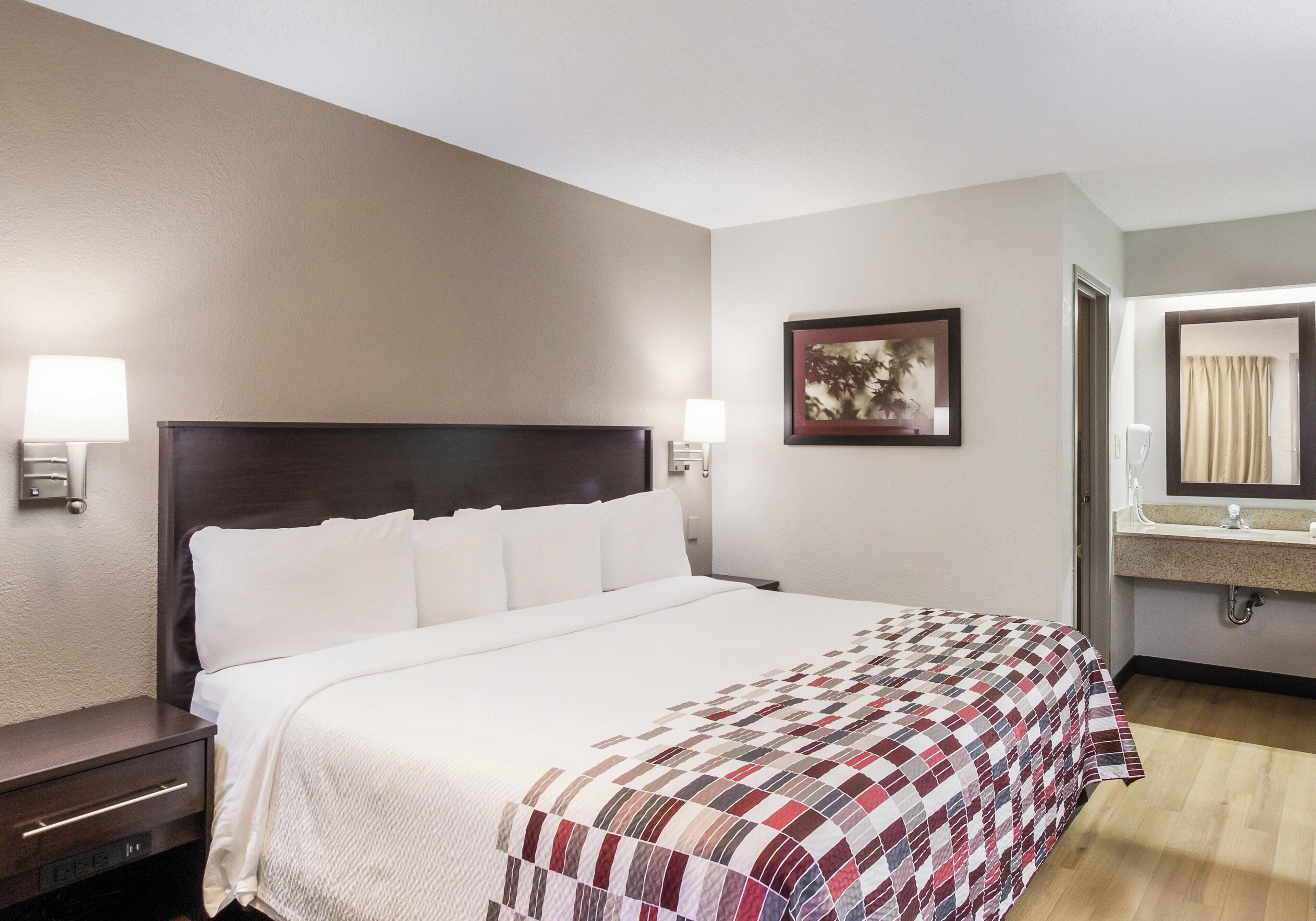 A hotel room with a bed, bedside tables, a nightstand, a wall-mounted lamp, a framed picture, and a bathroom with a sink and mirror.