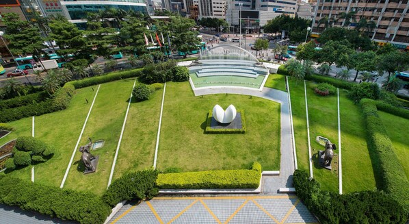 A landscaped public square with a central fountain, surrounded by neatly trimmed hedges and a few statues.