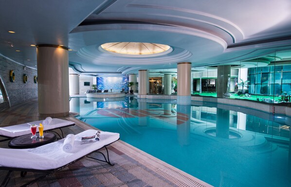 A spacious indoor swimming pool with lounge chairs, a central column, and a modern ceiling design.