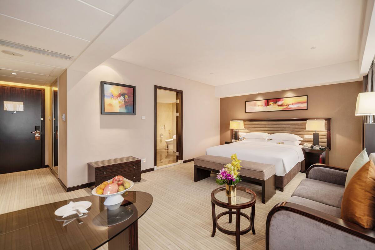 Una camera d'albergo con un letto, un divano, un tavolino con un vaso di fiori, una ciotola di frutta e una scrivania con un telefono.