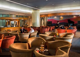Amplia sala de estar con diversas opciones de asiento, un bar con cantinero y un piano al fondo.