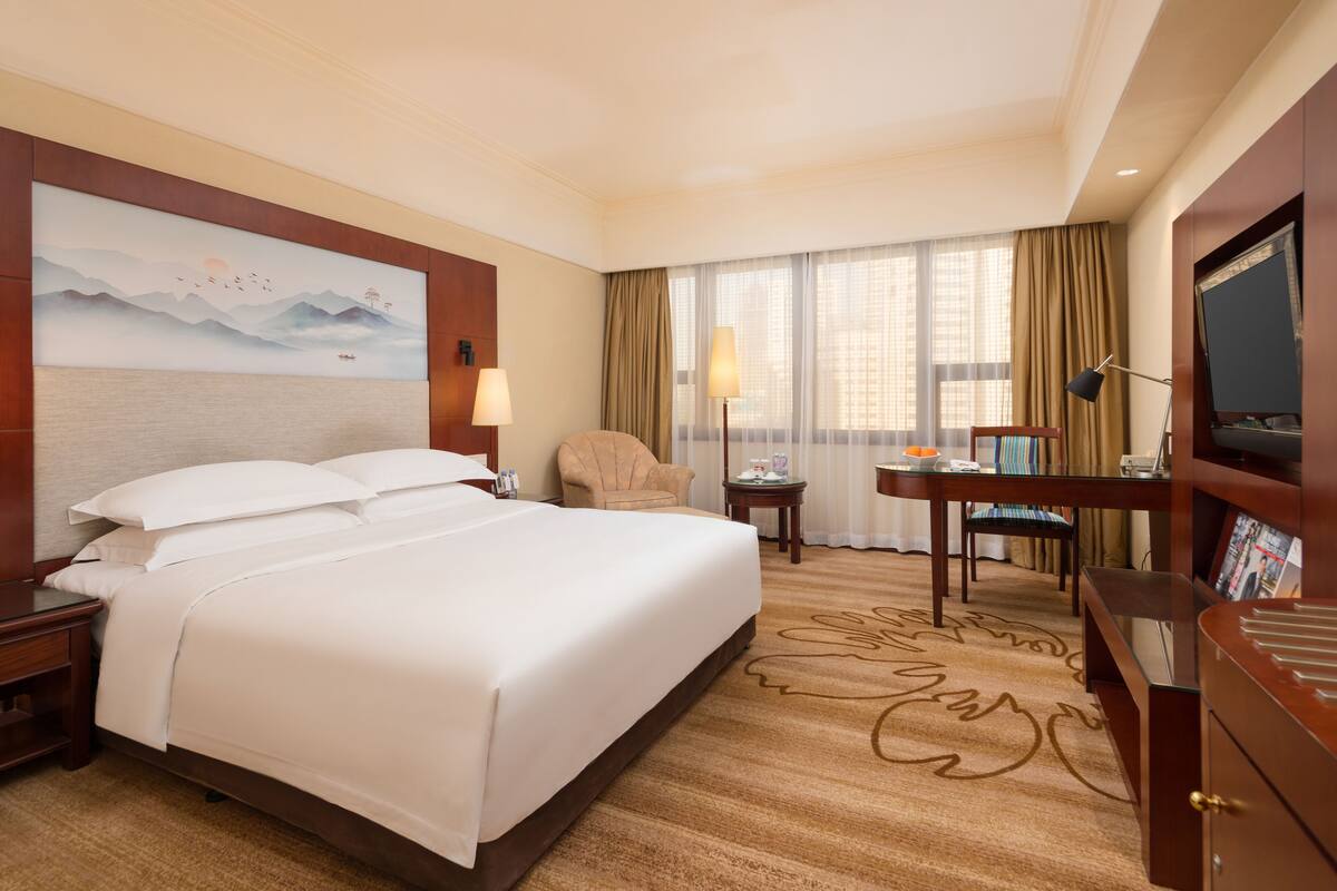 A hotel room with a large bed, a desk, a chair, a TV, and a painting on the wall.