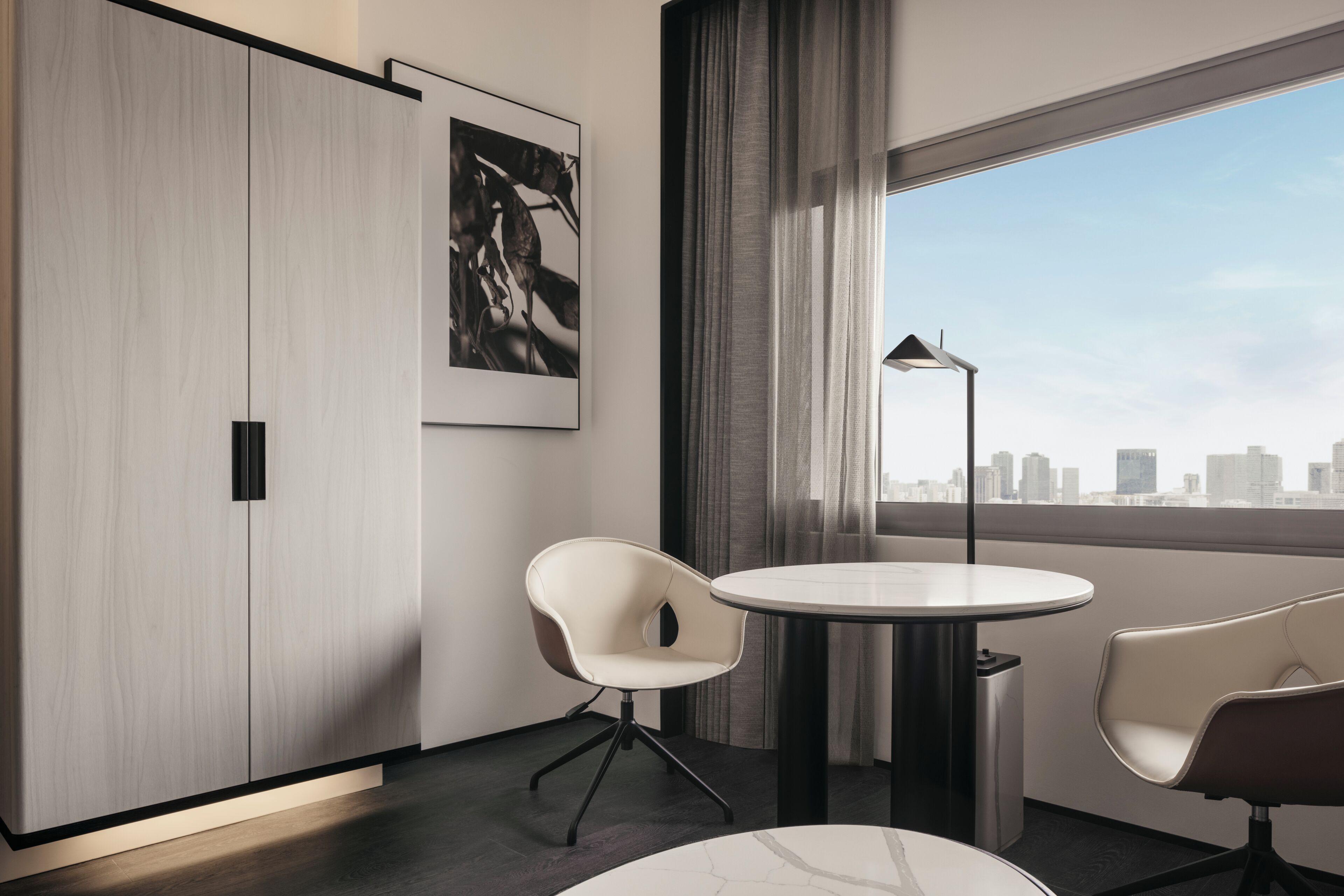 A modern hotel room with a large wardrobe, a round table, two chairs, a floor lamp, and a city view through a large window.