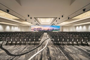 Un salón de conferencias amplio con filas de sillas, un escenario central y un mural con vistas a la ciudad en la pared de fondo.