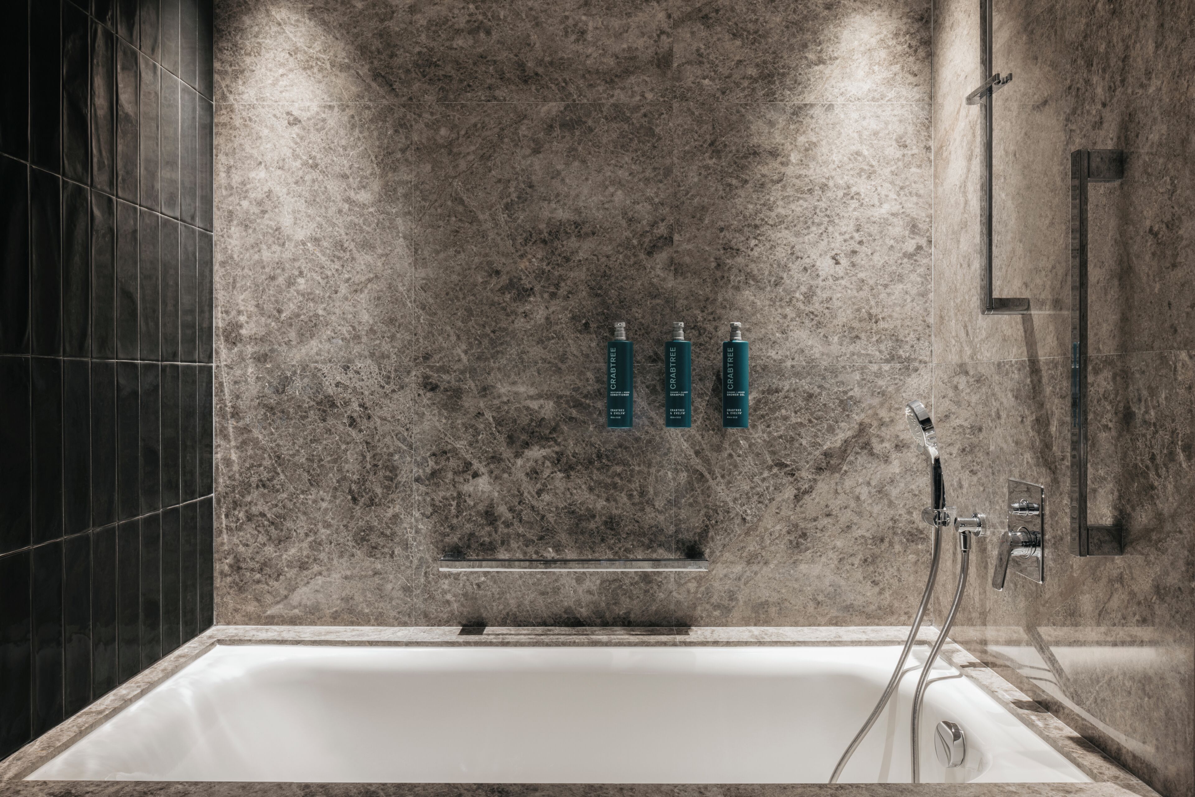 Une salle de bain moderne avec un mur en marbre, une baignoire et trois bouteilles de shampoing.