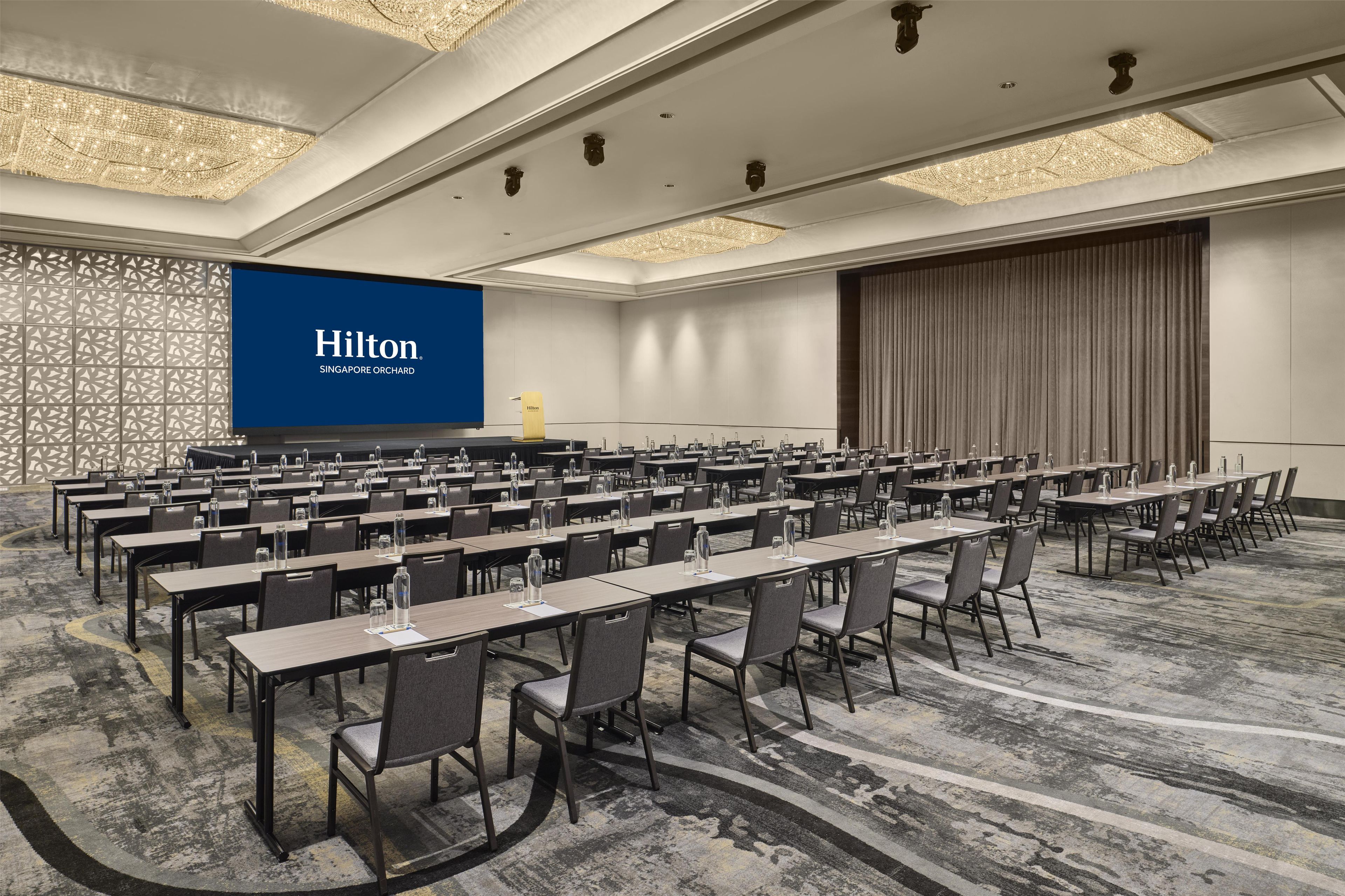 A conference room with multiple tables and chairs, a large screen displaying the Hilton logo, and a patterned carpet.
