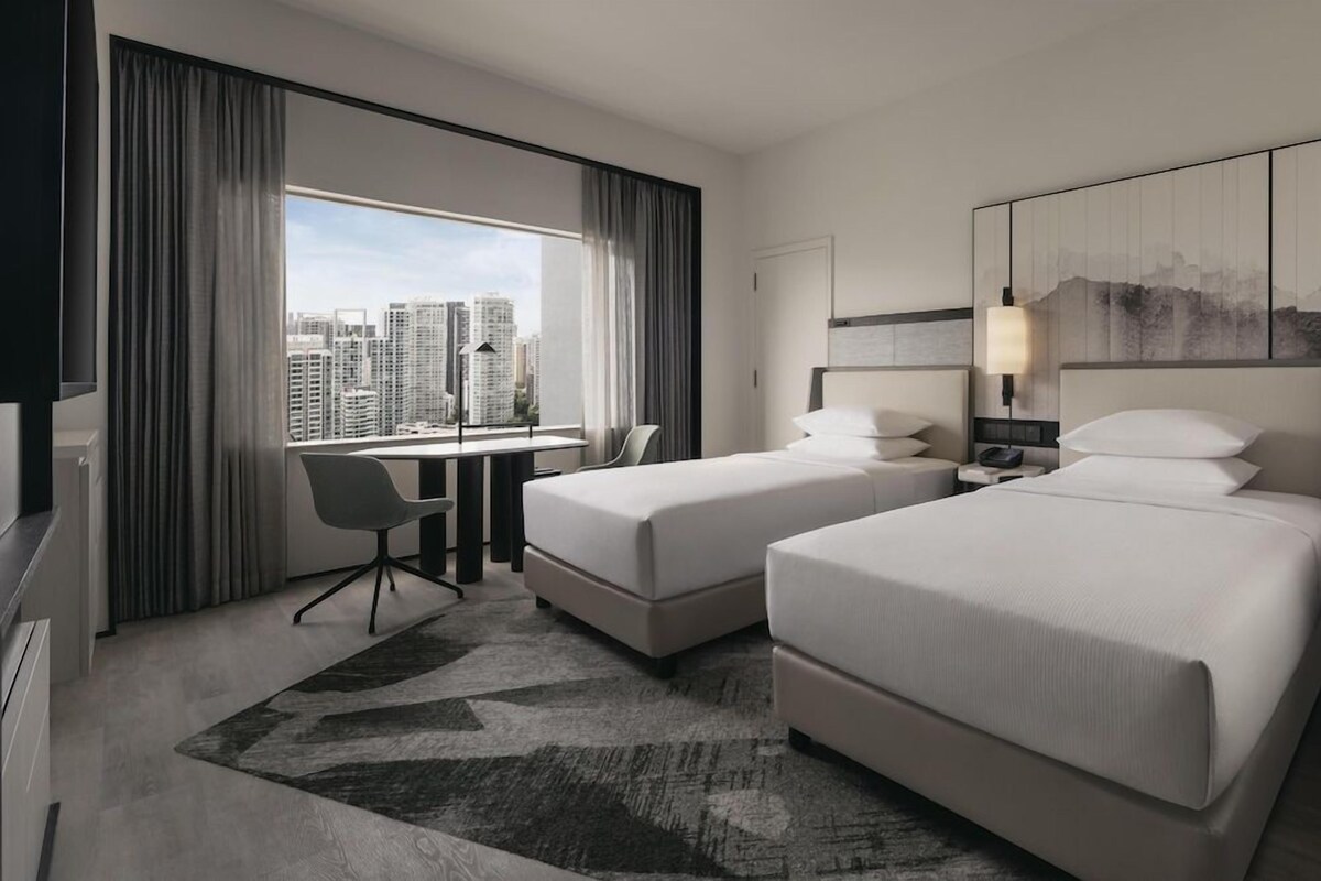 A hotel room with two beds, a desk, a chair, a large window with curtains, and a city view.