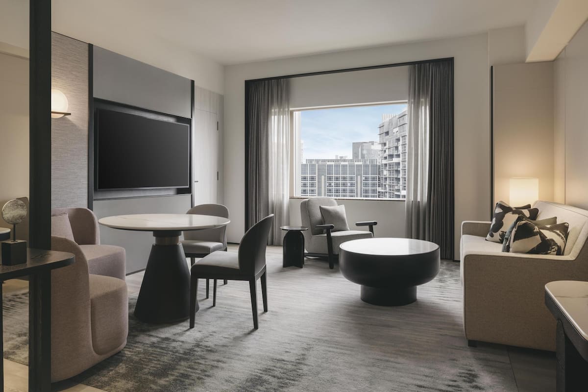 Una habitación de hotel moderna con televisor de pantalla plana en la pared, una mesa de centro redonda, un sofá, una mesa de comedor con sillas y un ventanal con cortinas.
