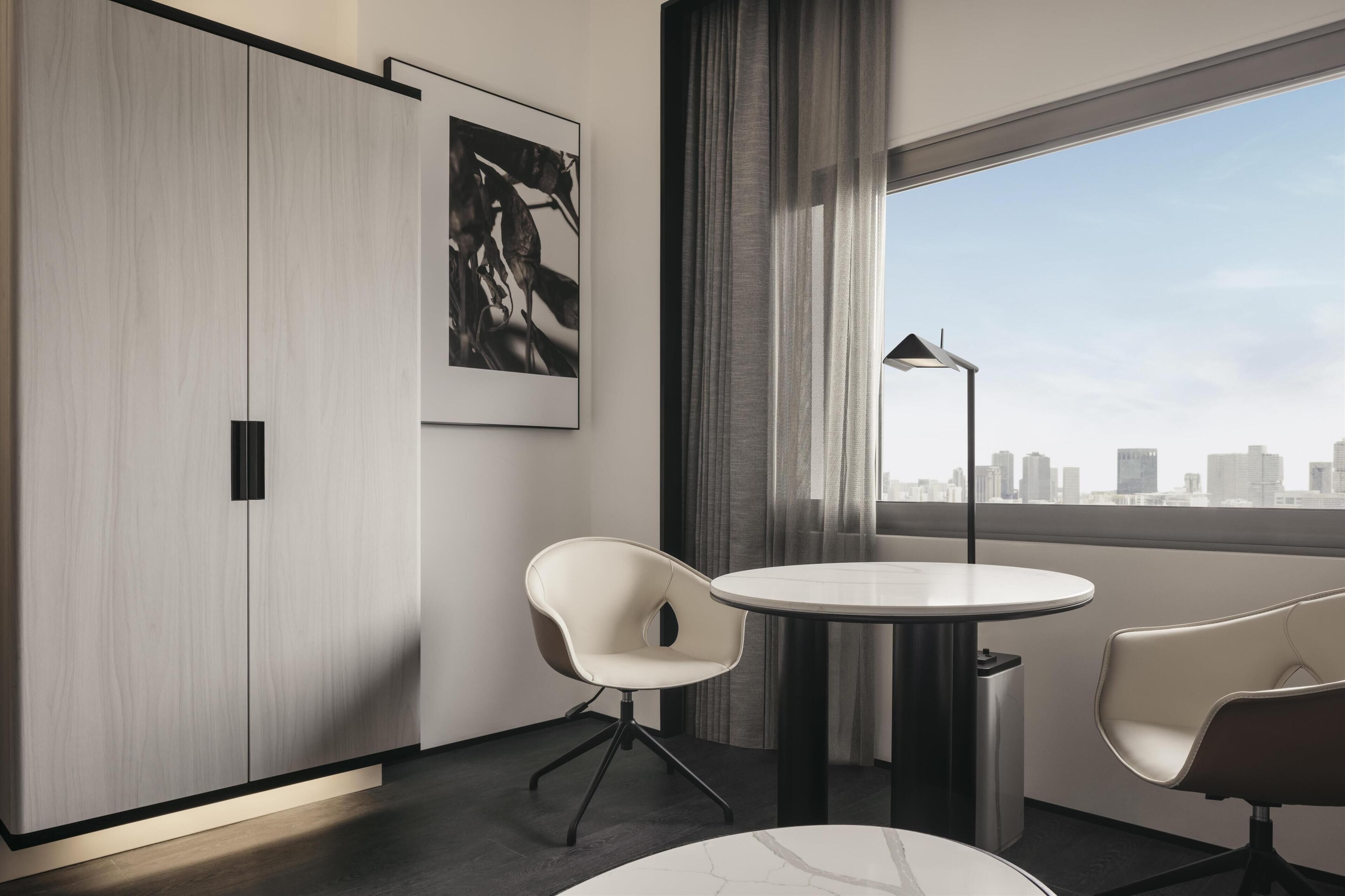 A modern hotel room with a large window, a round table, two chairs, a floor lamp, and a city view.