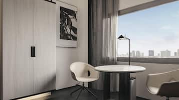 A modern hotel room with a large window, a round table, two chairs, a floor lamp, and a city view.