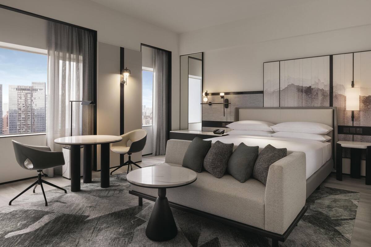 Habitación de hotel moderna con una cama grande, un sofá, una mesa de comedor y una silla. Se aprecia una vista de la ciudad a través de la ventana, y el diseño de la habitación es minimalista, con un espejo grande y obras de arte en la pared.