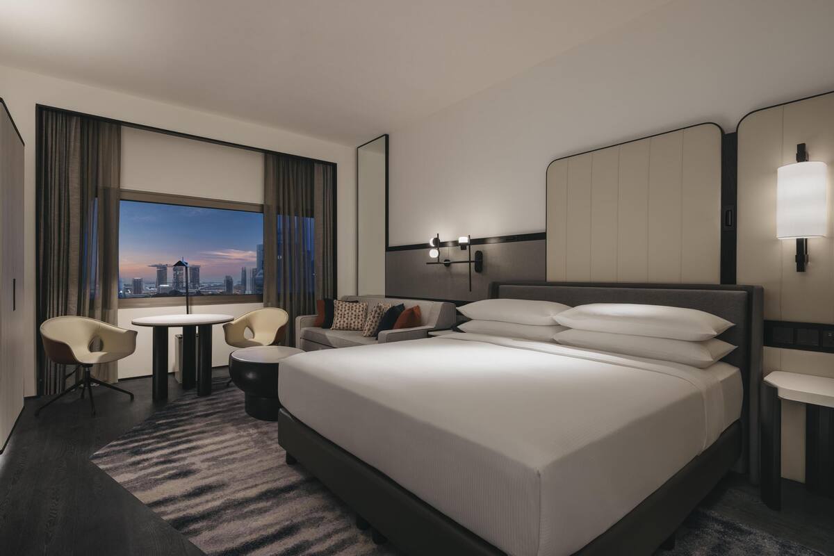Una habitación de hotel moderna con una cama grande, un escritorio, dos sillas, una mesita y vistas a la ciudad.