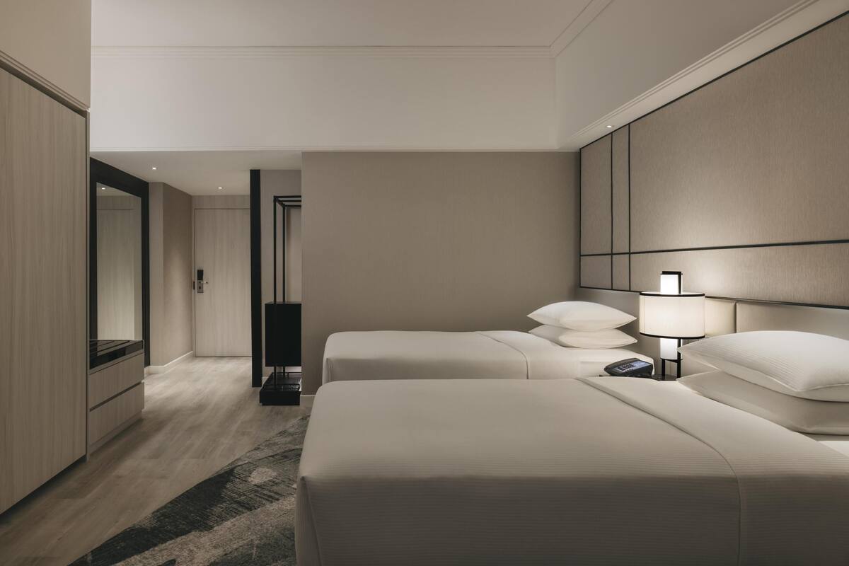 Una habitación de hotel moderna con dos camas, un espejo grande, un televisor de pantalla plana y una mesita de noche con lámpara.