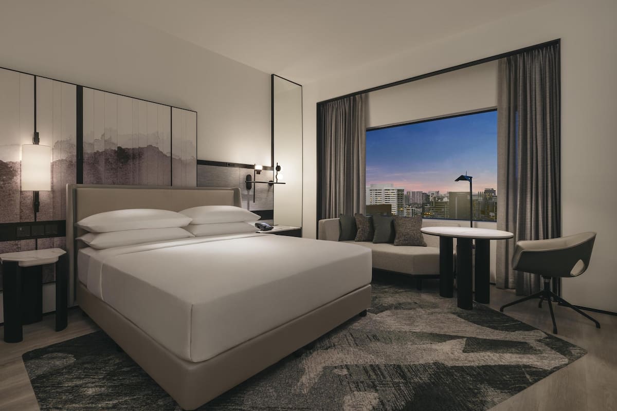 Una habitación de hotel moderna con una cama grande, un sofá, una mesita y vistas a la ciudad a través de un ventanal amplio.