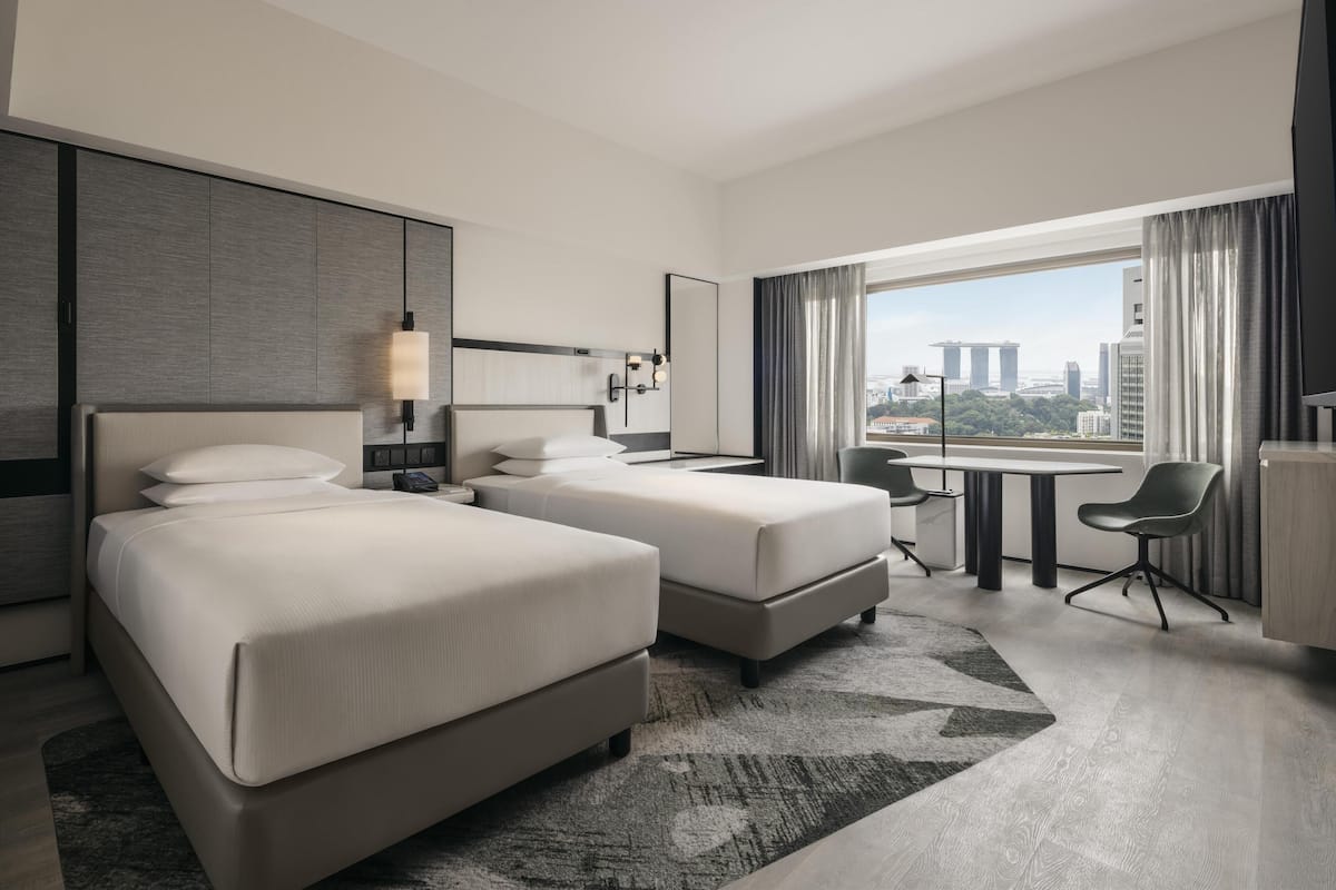 Habitación de hotel con dos camas, un escritorio, una silla y un ventanal con vista a la ciudad.