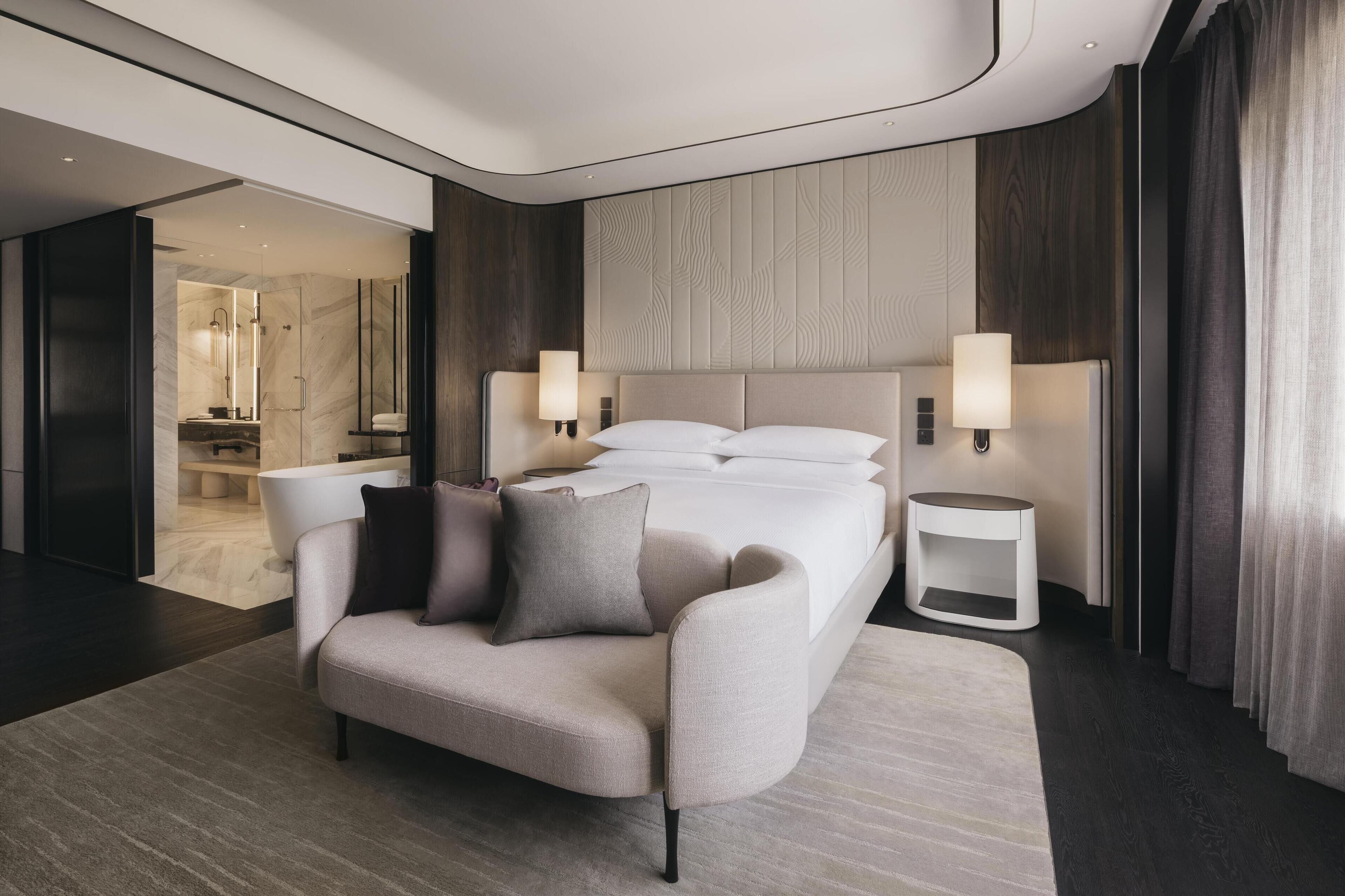 Una habitación de hotel moderna con una cama grande, dos lámparas de noche, un sofá y un baño visible a través de una puerta abierta.