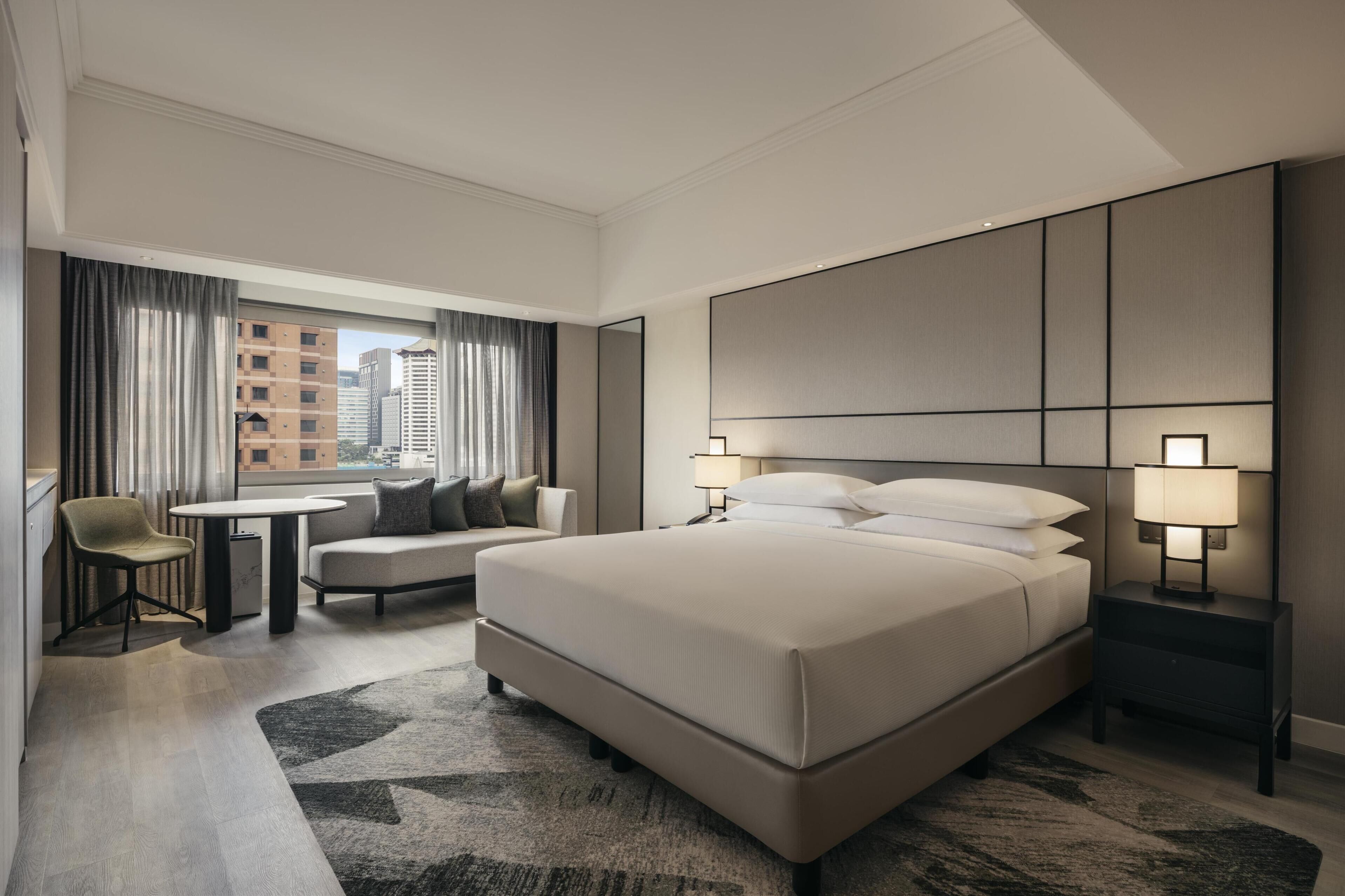 Una habitación de hotel moderna con una cama grande, una zona de estar con una mesa y sillas, y vista a la ciudad a través del ventanal.