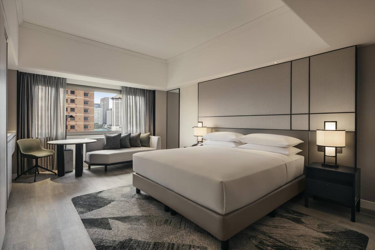 Una habitación de hotel moderna con una cama grande, una zona de estar con una mesa y sillas, y vista a la ciudad a través del ventanal.