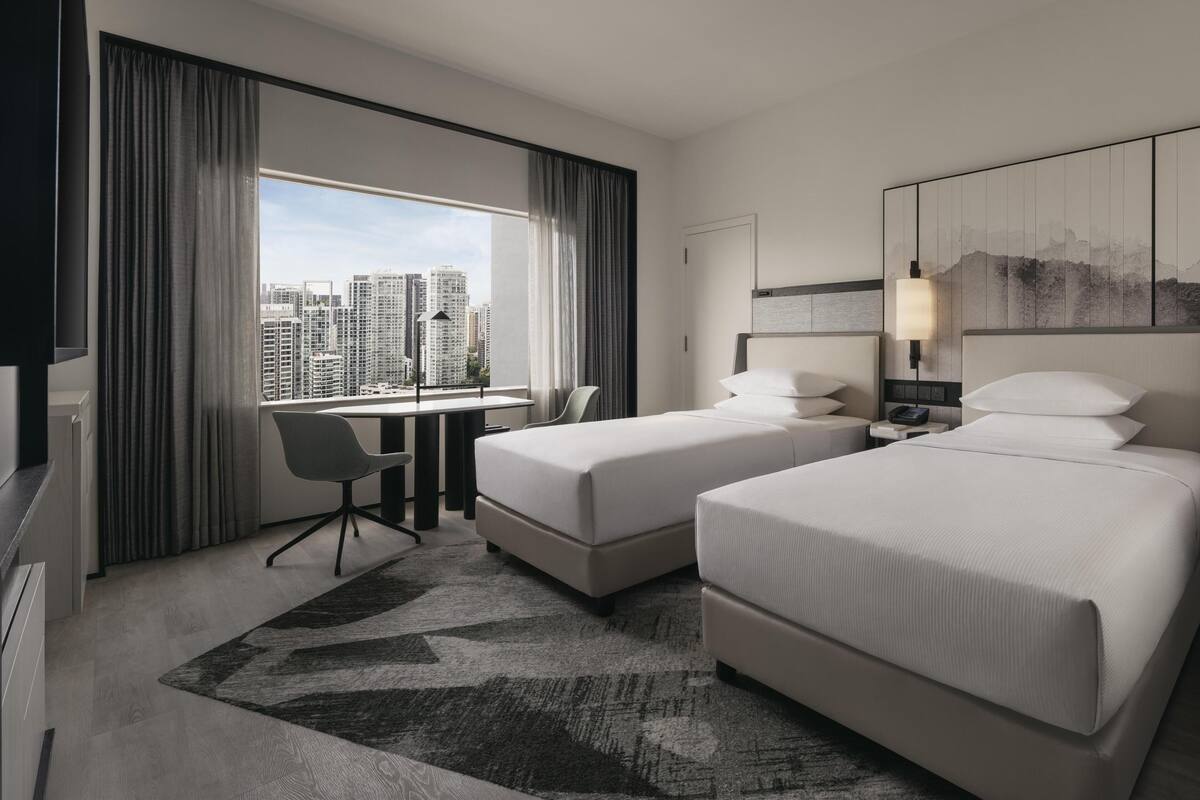Habitación de hotel con dos camas, un escritorio y una silla. Amplia ventana con cortinas, vista a la ciudad y una obra de arte colgada en la pared.