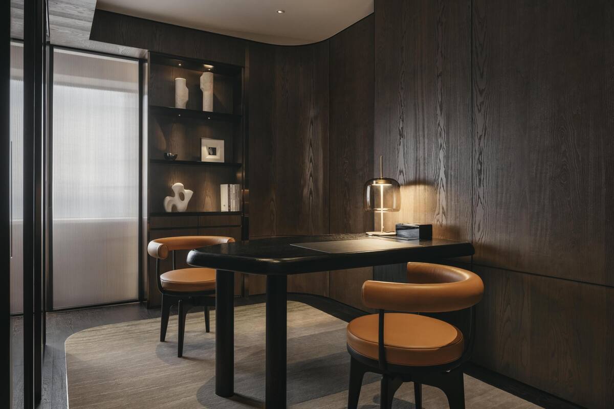 Un interior moderno con un panel de madera oscura, una mesa negra con dos sillas de cuero y una lámpara pequeña sobre la mesa.