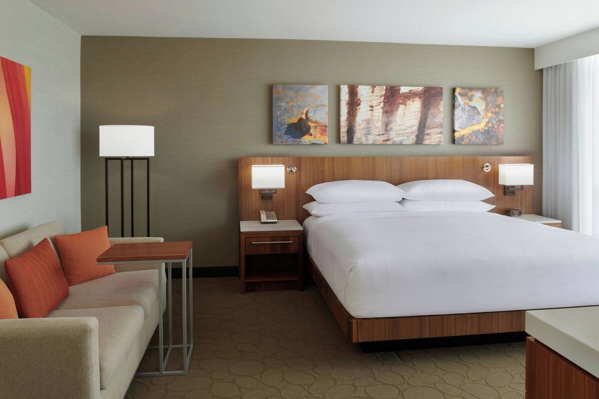 A hotel room with a large bed, a desk, a chair, and a nightstand with lamps. There are three paintings on the wall above the bed.