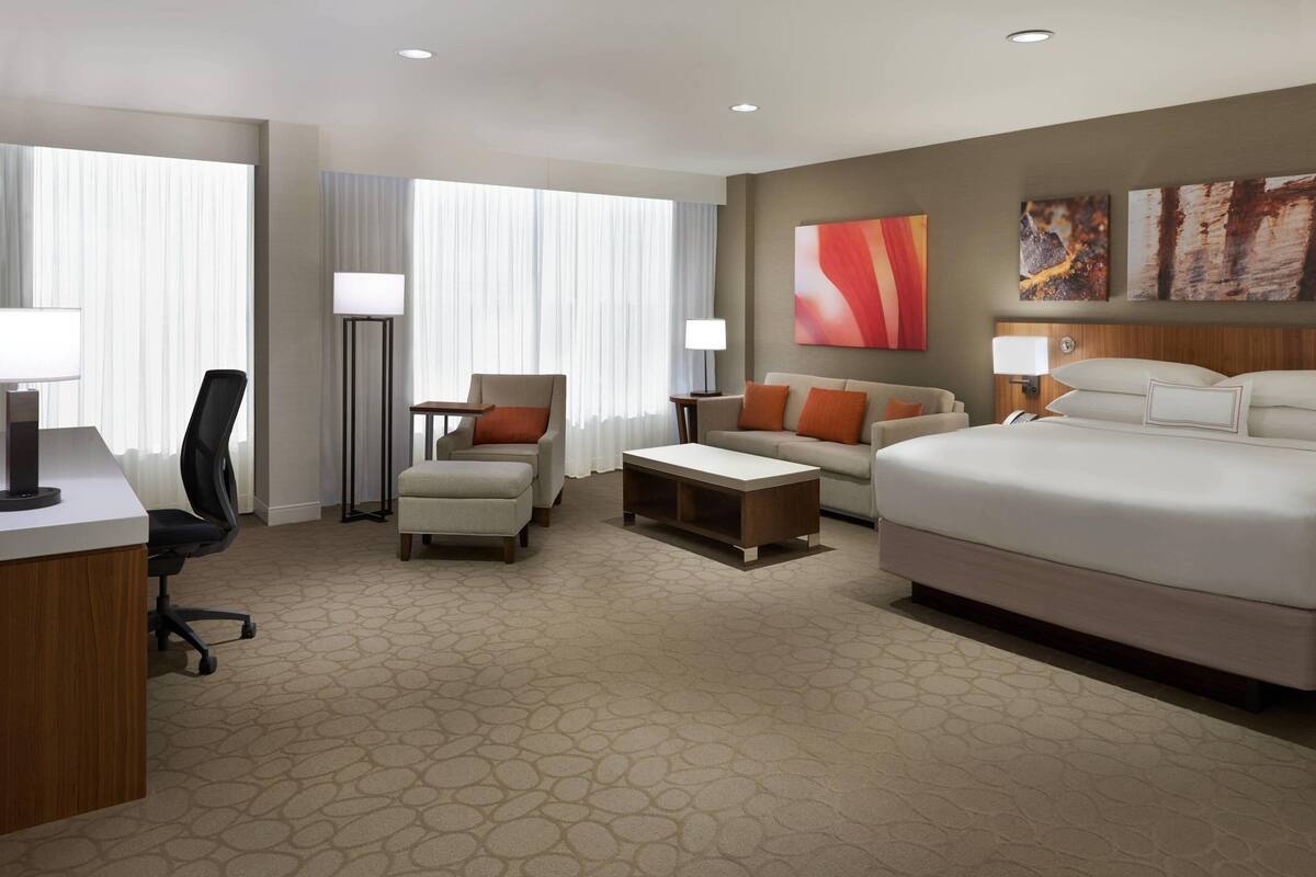 A hotel room with a bed, a sofa, a desk, and a chair. There are two lamps and a painting on the wall.