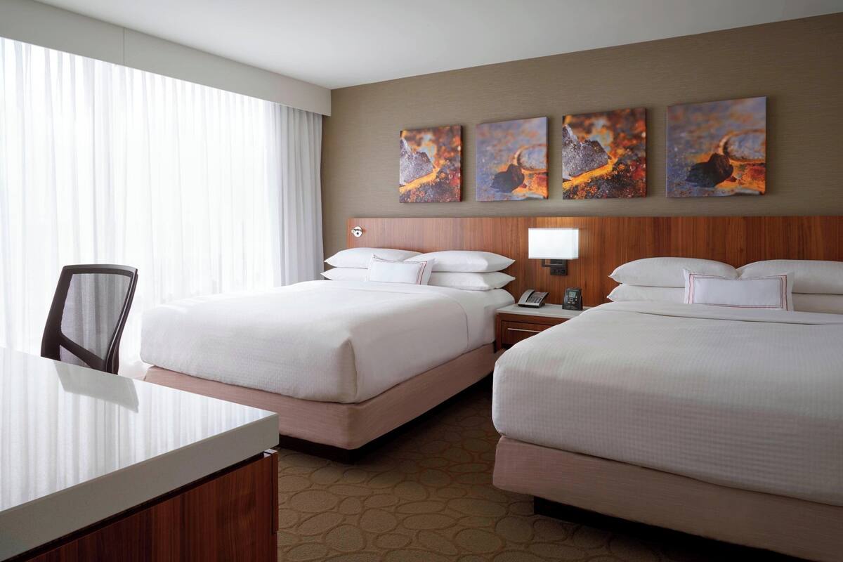 A hotel room with two beds, a desk, and a chair. There are three paintings on the wall above the beds.