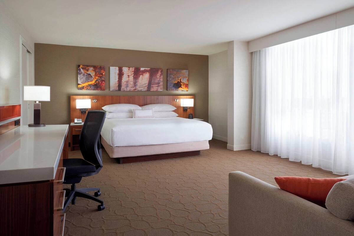 A hotel room with a bed, desk, chair, and large window with curtains.