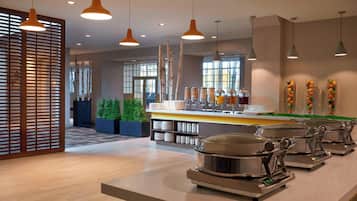Un buffet disposant de plusieurs points de service, d’un comptoir avec une variété de récipients contenant différents aliments, et d’un aménagement intérieur moderne avec un plancher en bois et des luminaires suspendus.