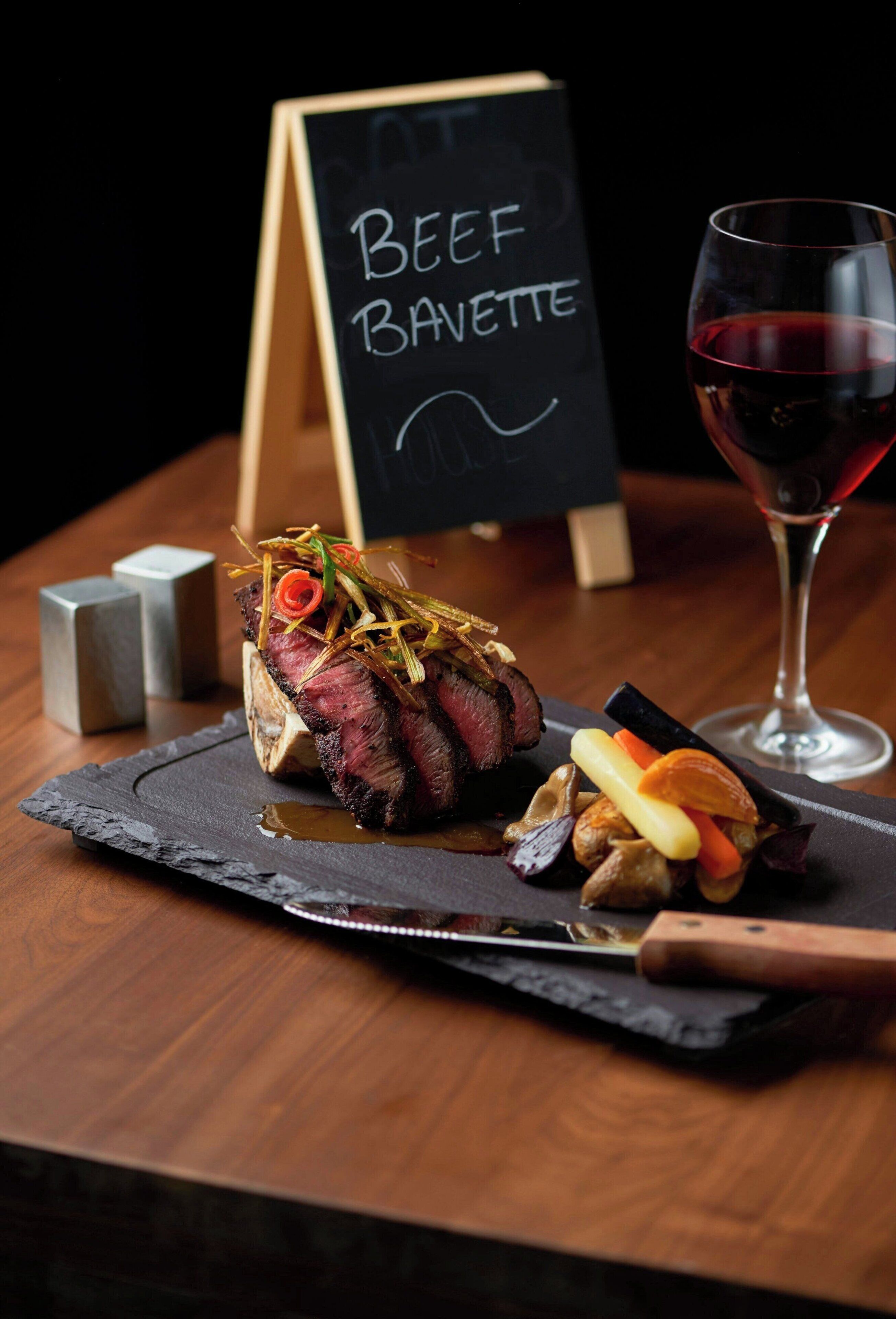Un plato de carne de res sobre una pizarra, con un cuchillo, una copa de vino tinto y un letrero de pizarra que dice 'Beef Bavette'.