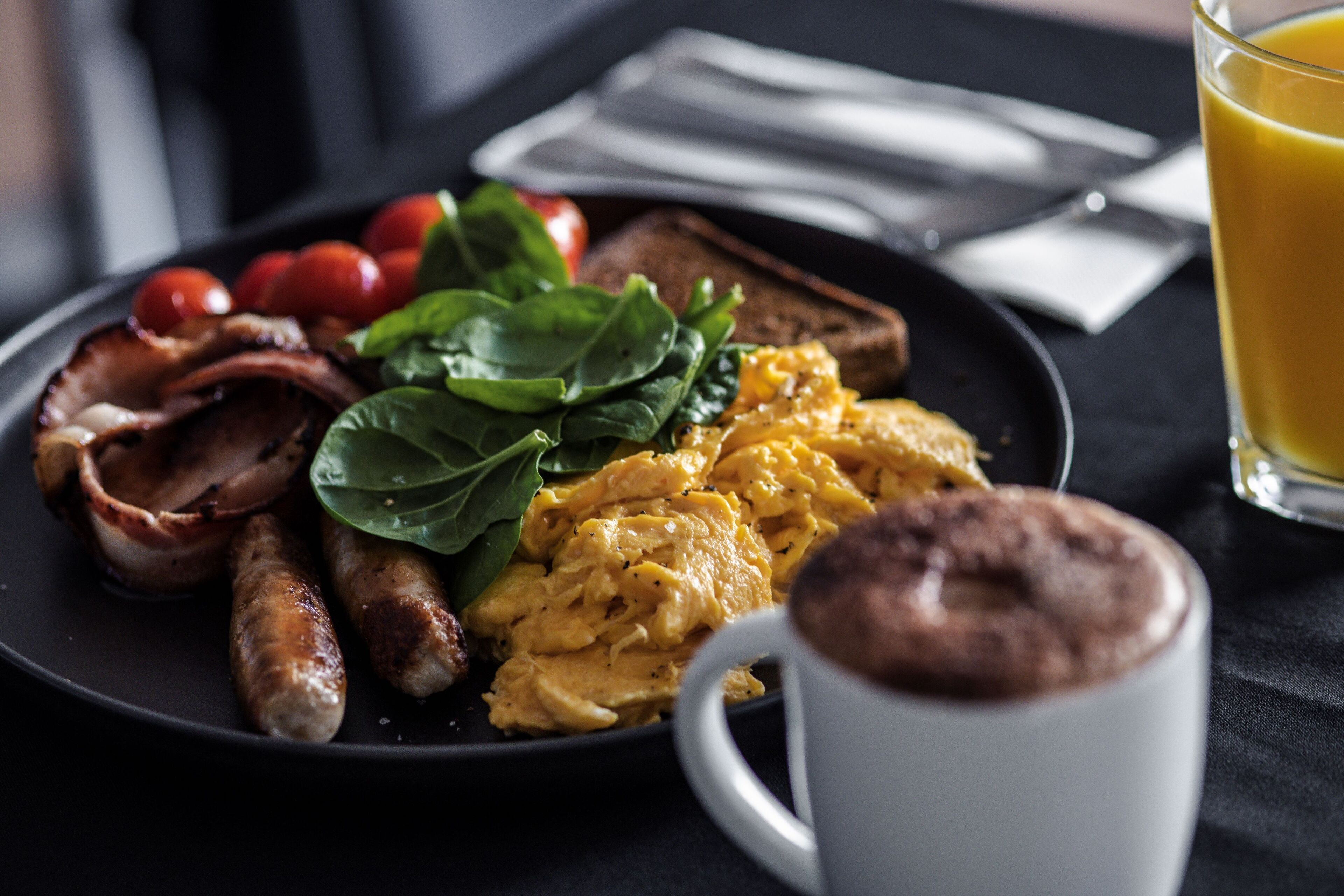 Un plato de desayuno con huevos, tocino, salchichas, tomates, espinacas, tostadas y un vaso de jugo de naranja.