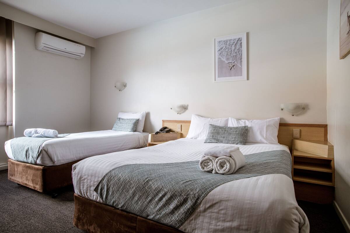 A hotel room with two beds, each with a wooden headboard, a bedside table, and a wall-mounted air conditioner.