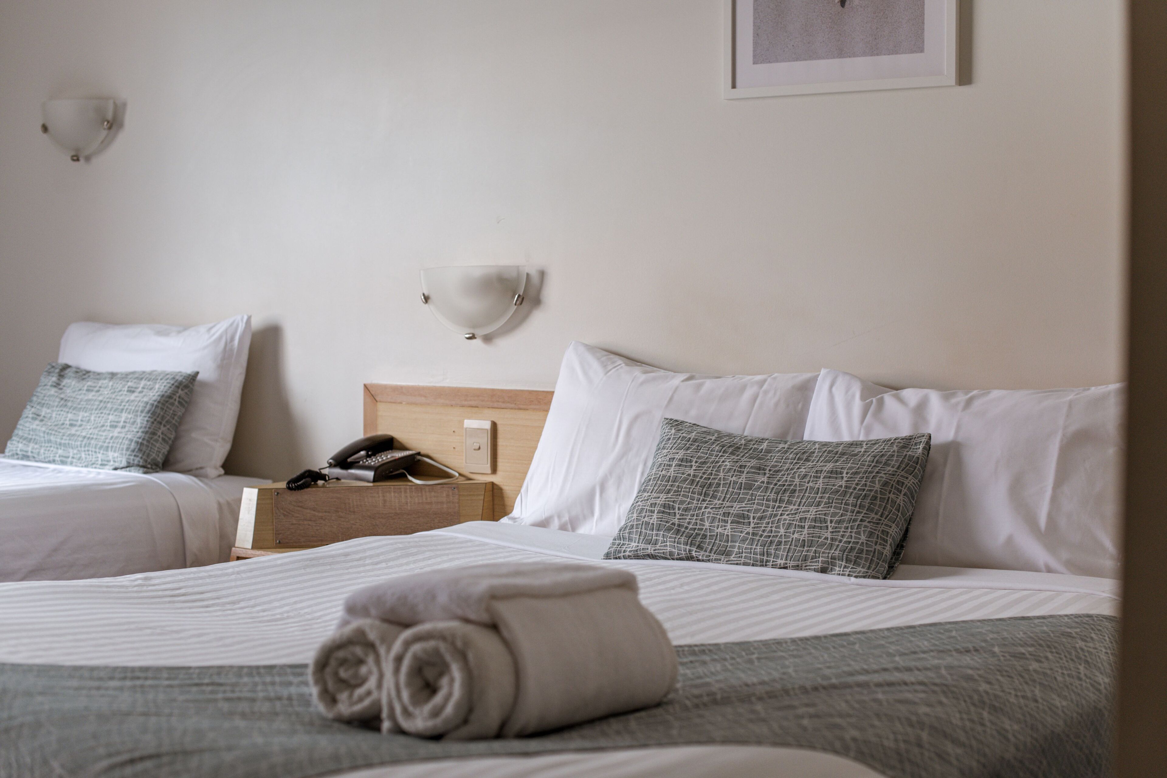 Ein Hotelzimmer mit zwei Betten, beide mit weißen Laken und gemusterten Kissen. Zwischen den Betten steht ein Nachttisch aus Holz mit einem Telefon und einer Lampe.