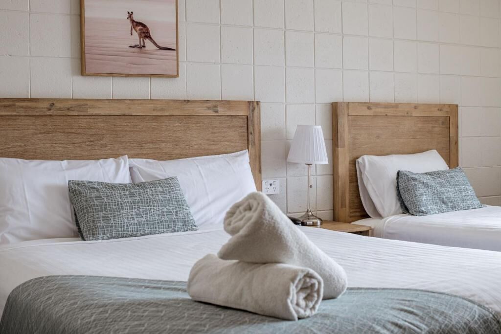 A hotel room with two beds, a wooden headboard, white bedding, a grey blanket, a grey pillow, a lamp, and a framed picture on the wall.