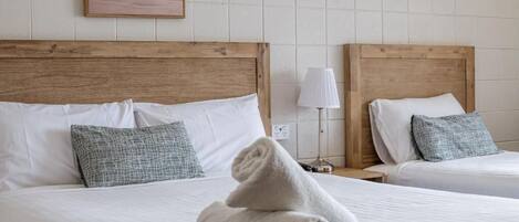 A hotel room with two beds, a wooden headboard, white bedding, a grey blanket, a grey pillow, a lamp, and a framed picture on the wall.
