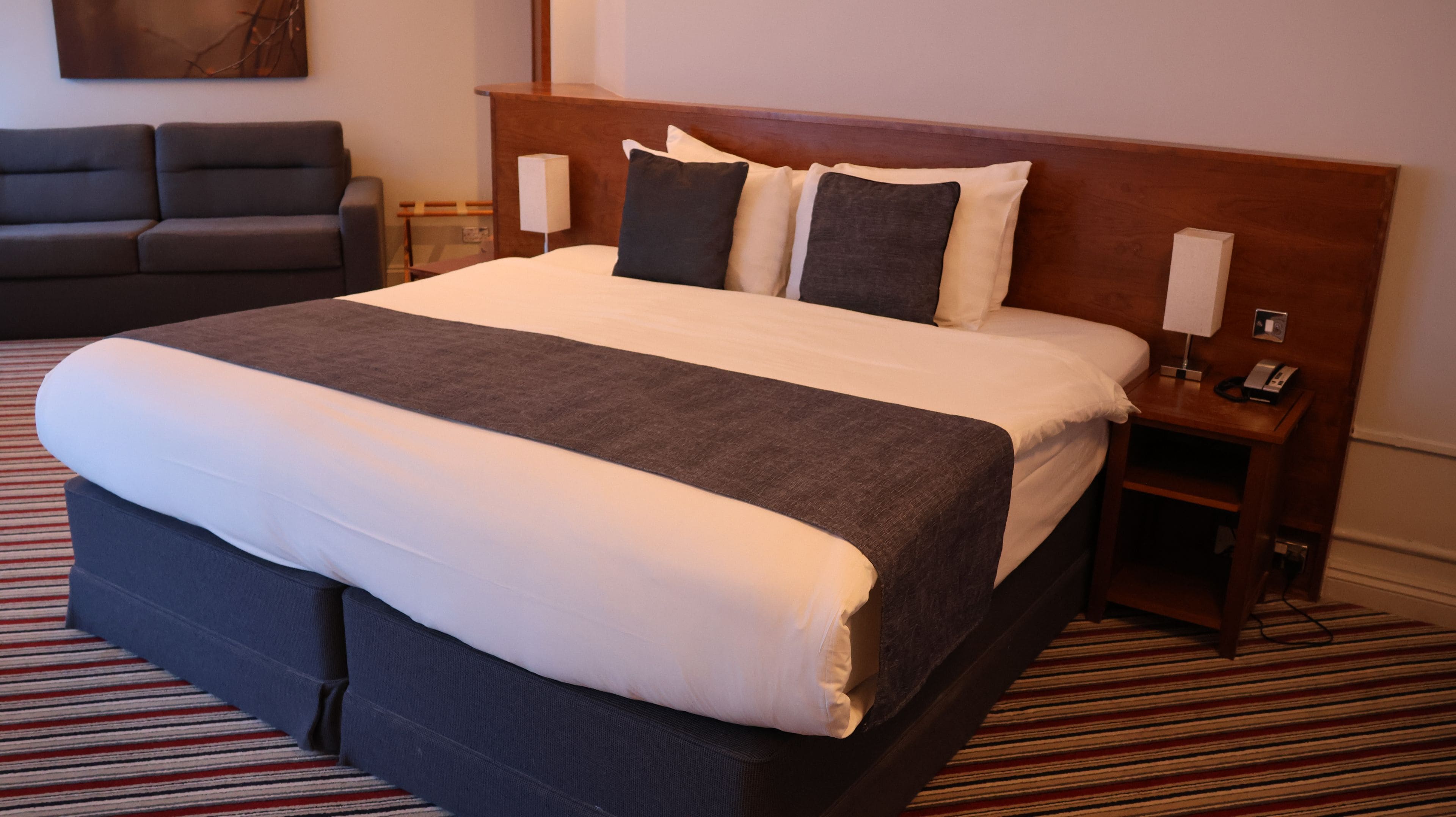 A hotel room with a large bed, a bedside table with a lamp, a small table, and a sofa.