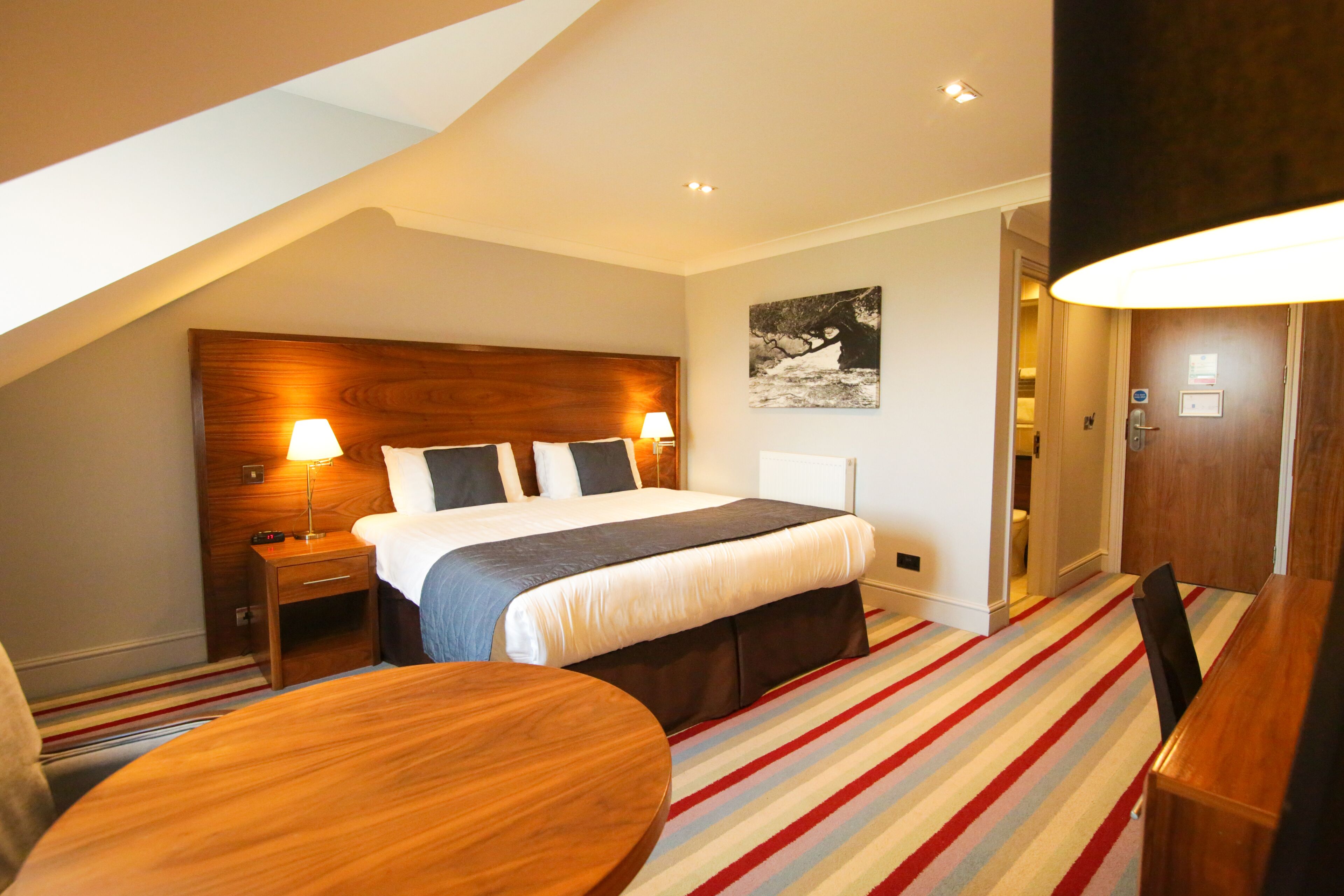A hotel room with a large bed, bedside tables with lamps, a striped carpet, and a wooden desk.